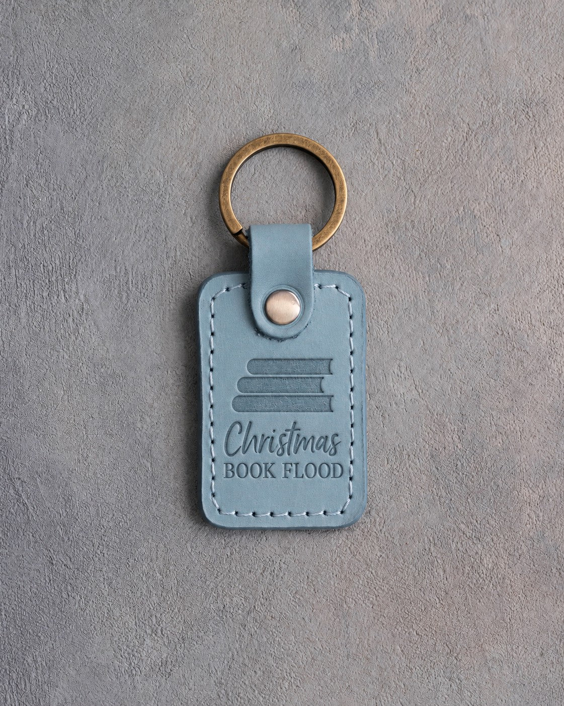 Turquoise leather keychain debossed with a stack of books above Christmas and BOOK FLOOD, on gray concrete