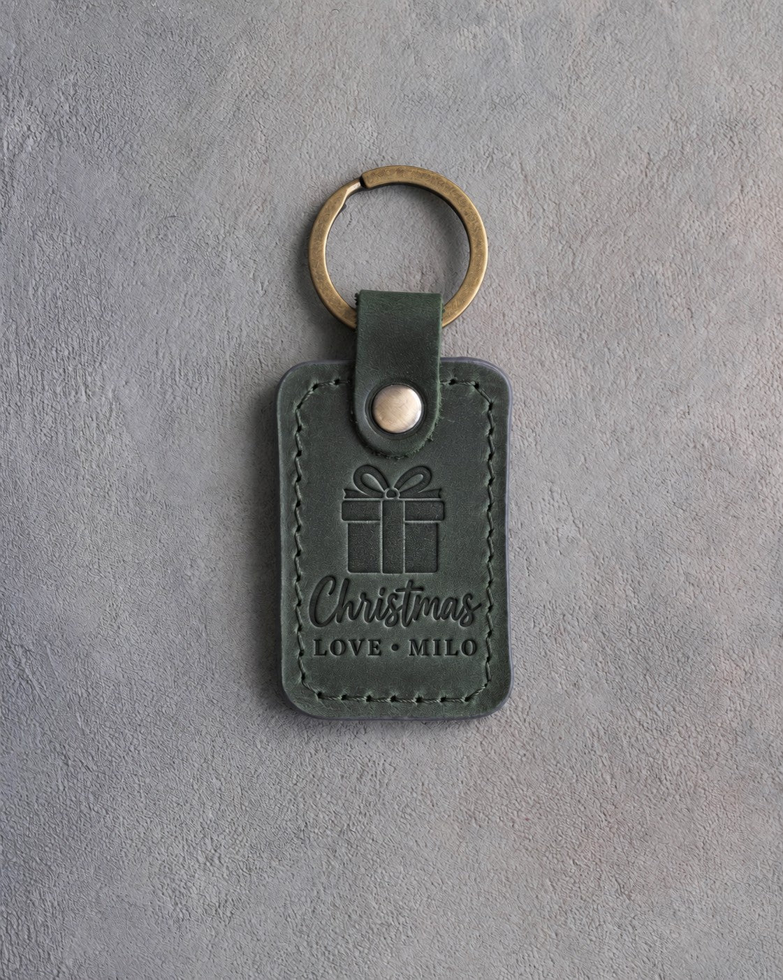 Forest green leather keychain debossed with a wrapped gift box above Christmas and LOVE · MILO, on gray concrete