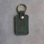 Forest green leather keychain debossed with a wrapped gift box above Merry Christmas and LOVE · THE DOG, on gray concrete