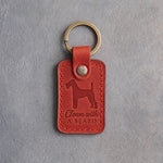 Poppy red leather keychain debossed with an Airedale terrier and the words Clown with A BEARD, on gray concrete