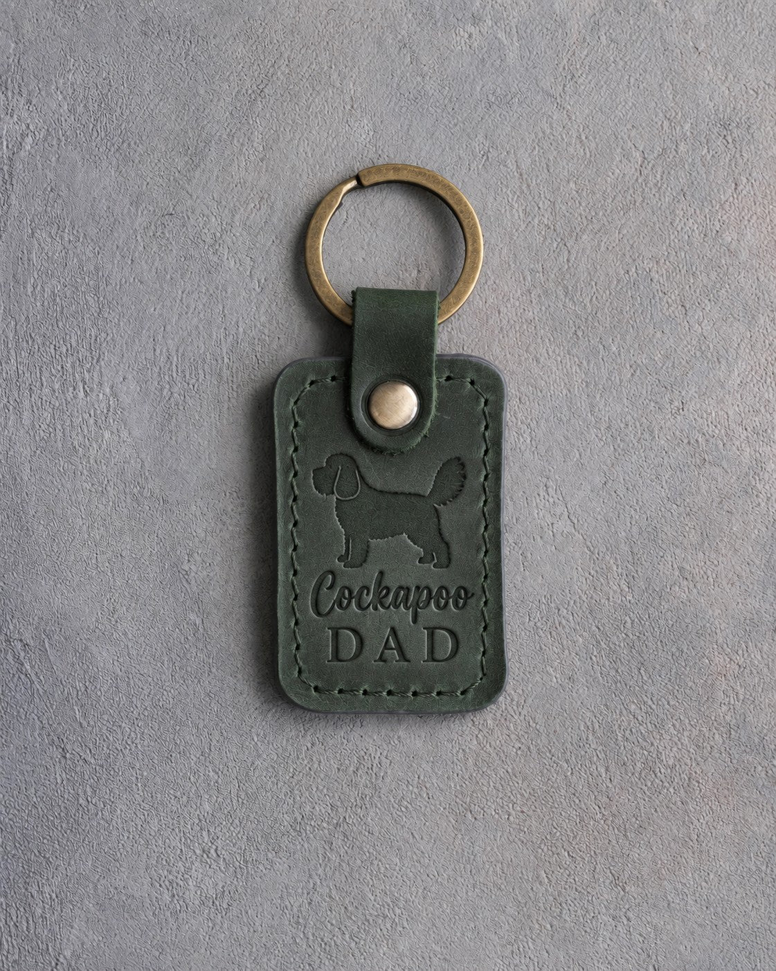 Forest green leather keychain debossed with a cockapoo silhouette and Cockapoo DAD on gray concrete