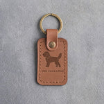 Caramel leather keychain debossed with a curly cockapoo above THE COCKAPOO, on gray concrete