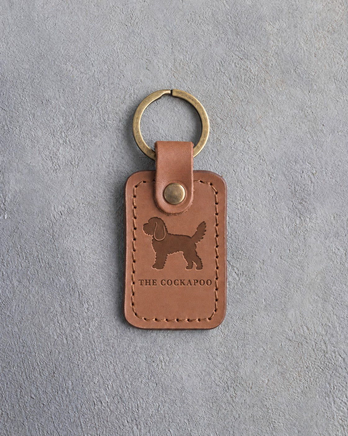 Caramel leather keychain debossed with a curly cockapoo above THE COCKAPOO, on gray concrete