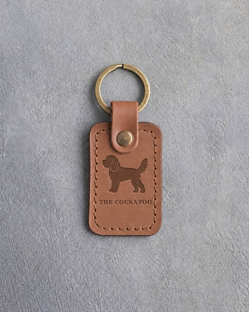 Caramel leather keychain debossed with a curly cockapoo above THE COCKAPOO, on gray concrete