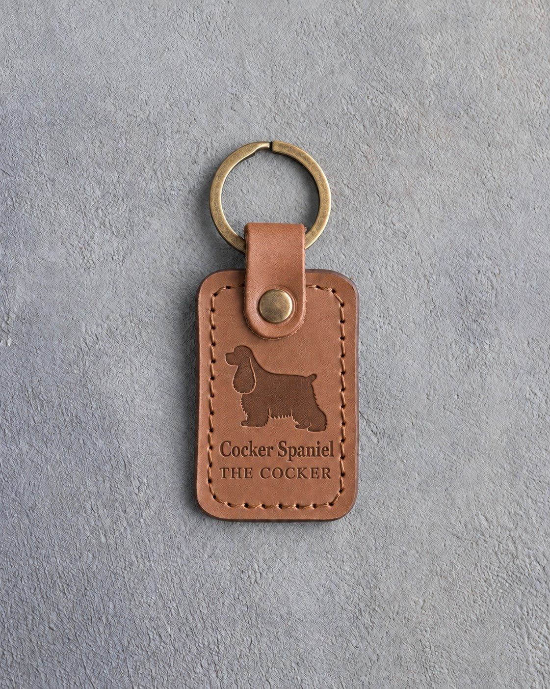Caramel leather keychain debossed with a long-eared cocker spaniel over Cocker Spaniel and THE COCKER, on gray concrete