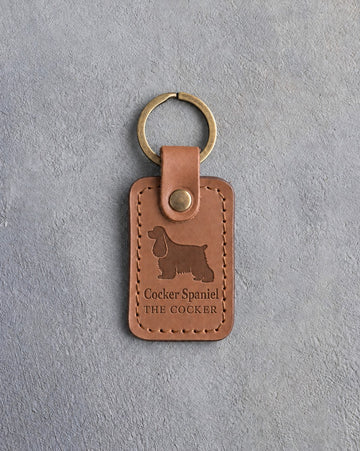 Caramel leather keychain debossed with a long-eared cocker spaniel over Cocker Spaniel and THE COCKER, on gray concrete