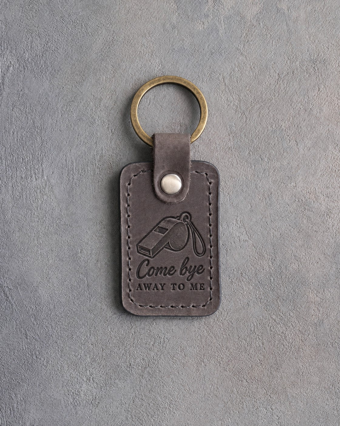 Country gray leather keychain debossed with a herding whistle on a lanyard and the words Come bye AWAY TO ME, on gray concrete