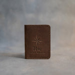 Sicilian brown leather card wallet standing on gray plaster, debossed with an eight-point compass rose above Wes and TRUE NORTH