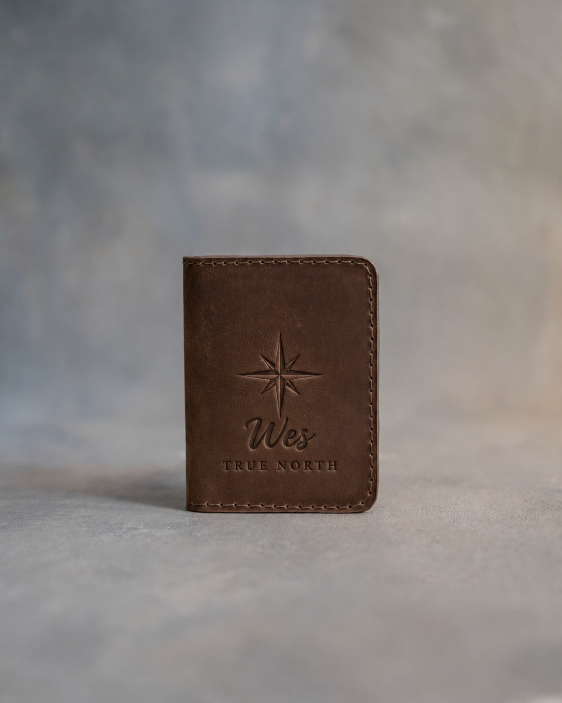Sicilian brown leather card wallet standing on gray plaster, debossed with an eight-point compass rose above Wes and TRUE NORTH