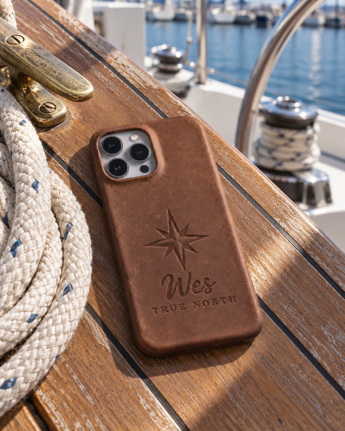 Sicilian brown Wes TRUE NORTH iPhone case in full-grain leather on the worn teak cockpit table of a sailboat beside a coiled white dock line