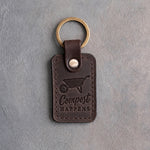 Dark espresso leather keychain debossed with a wheelbarrow above Compost HAPPENS, on gray concrete