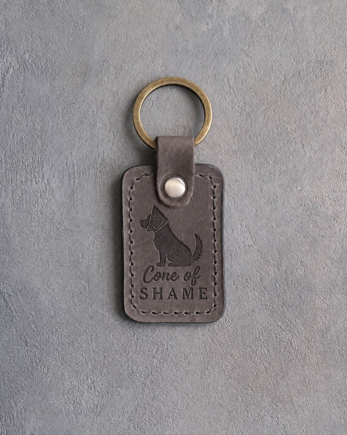Country gray leather keychain debossed with a sitting dog in a cone collar and the words Cone of SHAME, on gray concrete