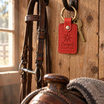 Poppy red Cowgirl SINCE 1998 keychain hanging from an iron bridle hook on rough pine boards in a tack room