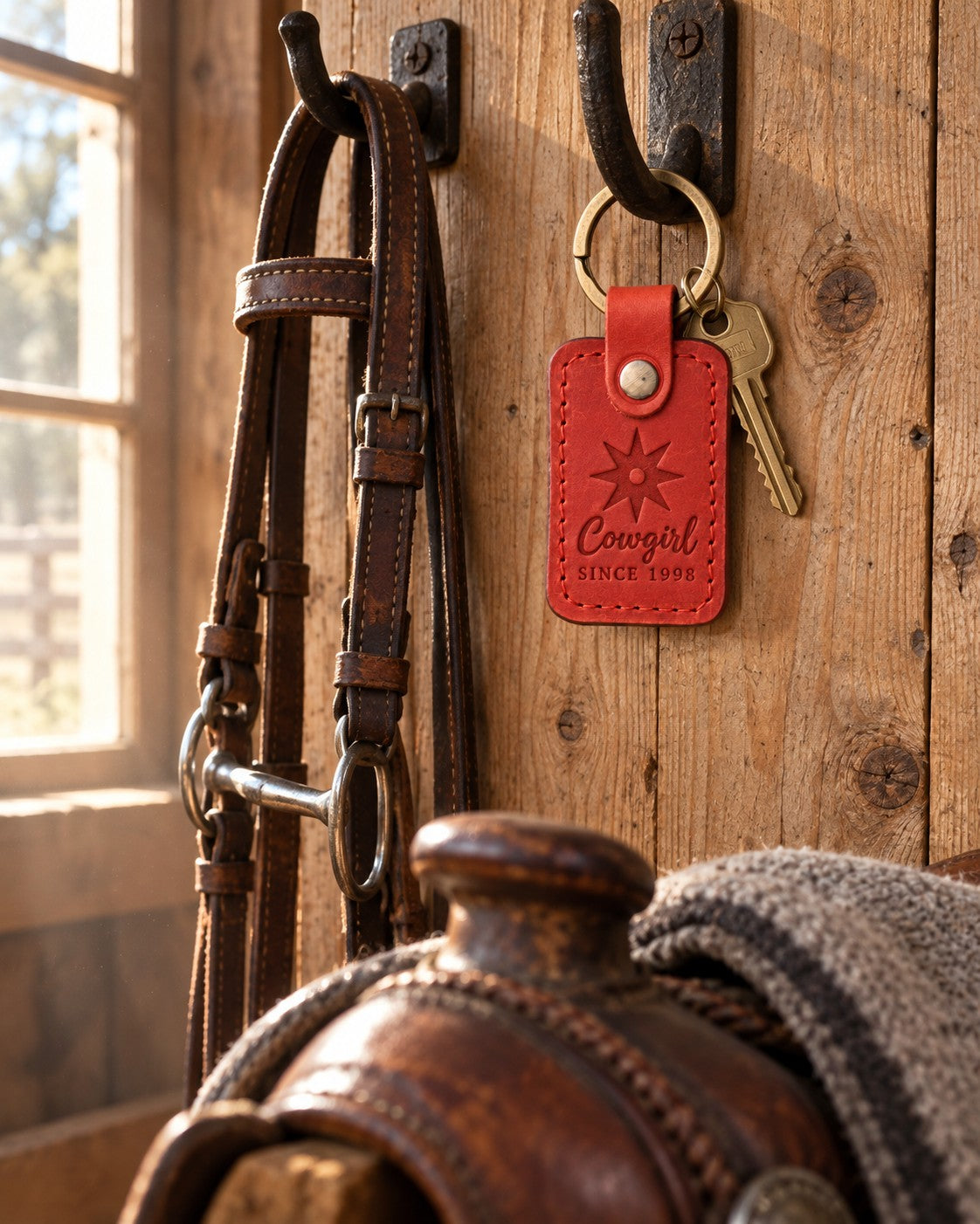 Poppy red Cowgirl SINCE 1998 keychain hanging from an iron bridle hook on rough pine boards in a tack room