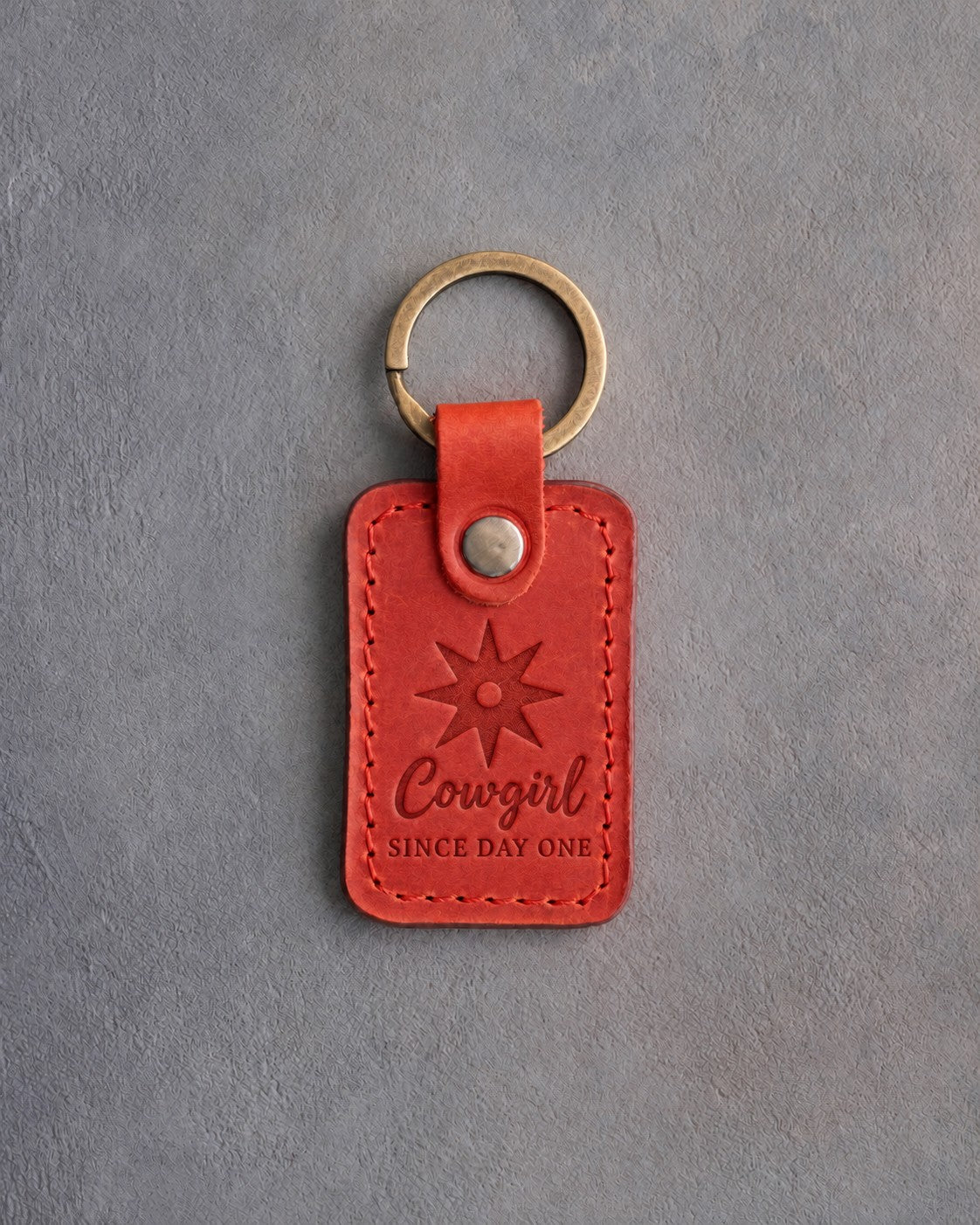 Poppy red leather keychain debossed with a spur rowel above Cowgirl and SINCE DAY ONE, on gray concrete