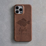 Back of a Sicilian brown leather iPhone case debossed with a crappie above Lyle and CRAPPIE below the camera, on gray plaster