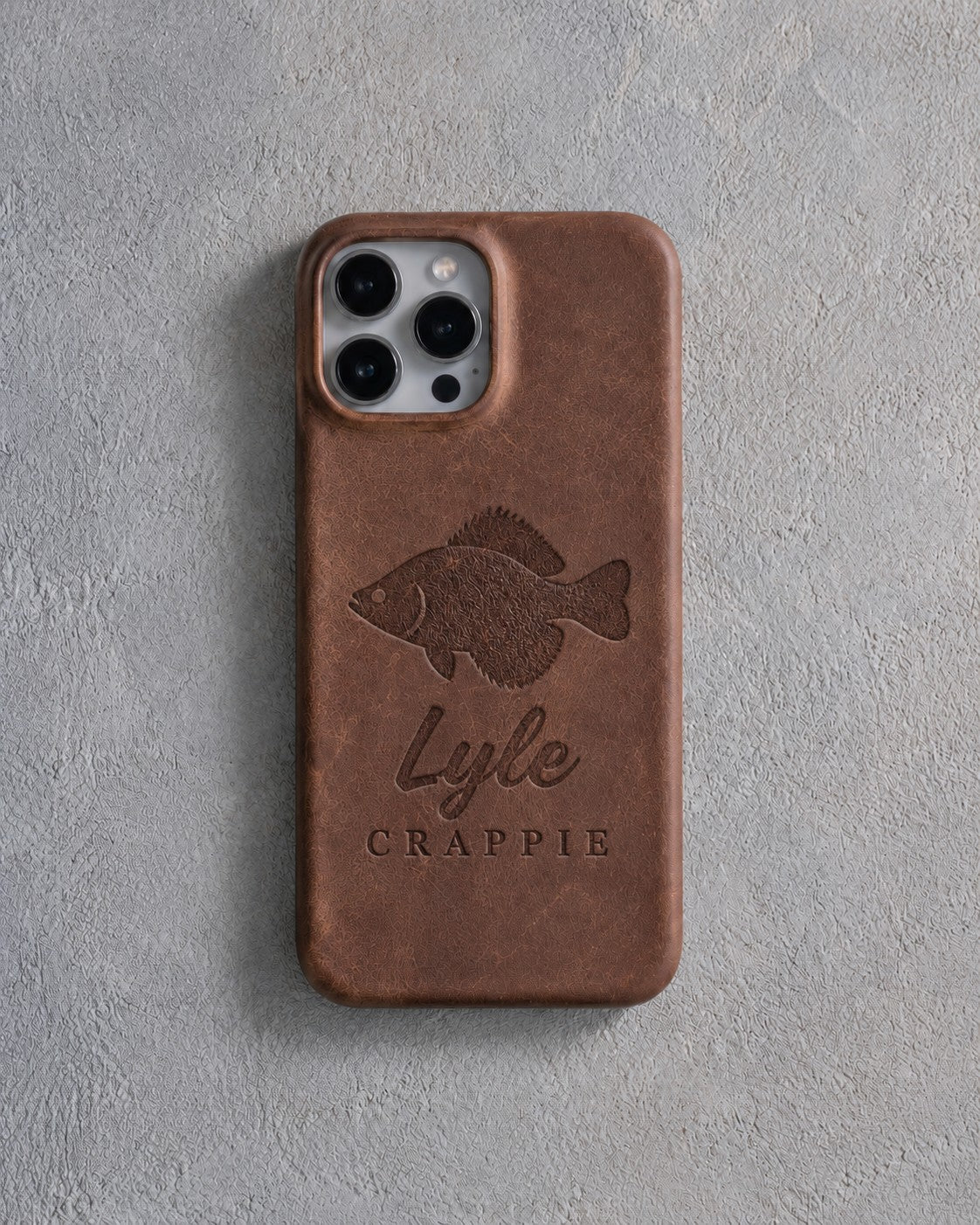 Back of a Sicilian brown leather iPhone case debossed with a crappie above Lyle and CRAPPIE below the camera, on gray plaster