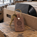 Crate Digger keychain in brown leather resting on the edge of a crate of used vinyl records in a record shop