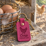 Bright pink Crazy Chicken Lady keychain on a weathered pine nesting box ledge beside brown eggs in a wire basket, a hen soft in the run behind