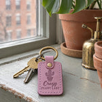 Lilac Crazy Plant Lady keychain on a windowsill crowded with small potted cacti and succulents