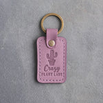 Lilac leather keychain debossed with a potted cactus above Crazy PLANT LADY, on gray concrete
