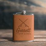 Caramel leather-wrapped hip flask debossed with crossed fishing rods above Garrett and TIGHT LINES, on a wooden table
