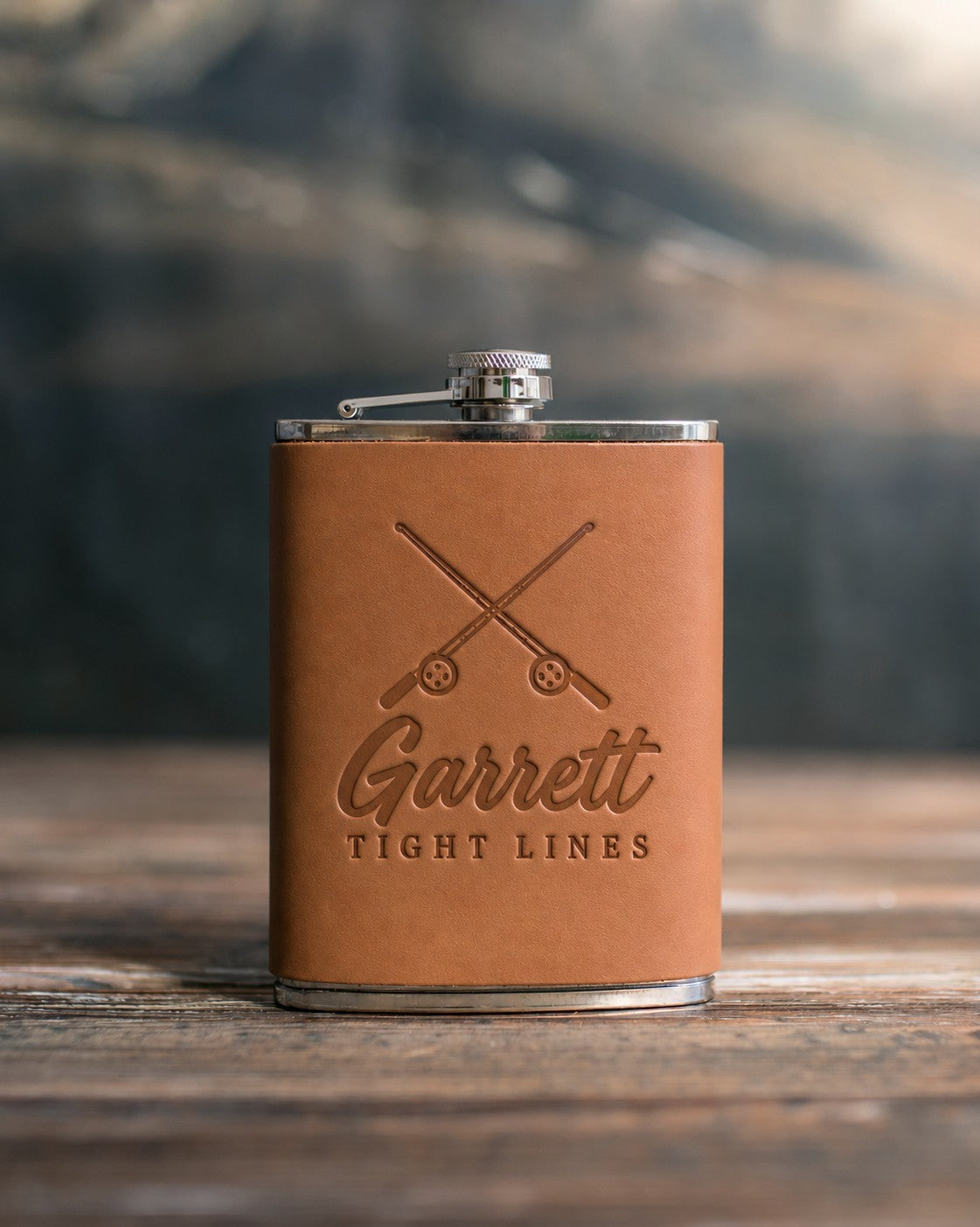Caramel leather-wrapped hip flask debossed with crossed fishing rods above Garrett and TIGHT LINES, on a wooden table