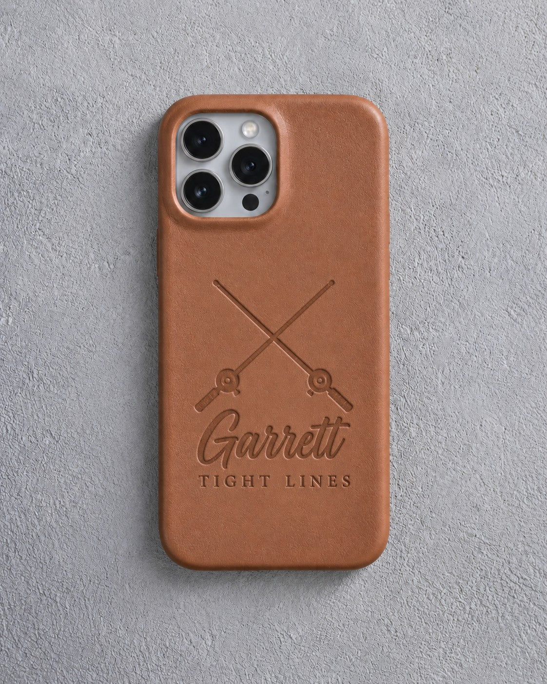Back of a caramel leather iPhone case debossed with crossed fishing rods above Garrett and TIGHT LINES below the camera, on gray plaster
