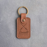 Caramel leather keychain debossed with crossed fishing rods above Garrett and TIGHT LINES, on gray plaster