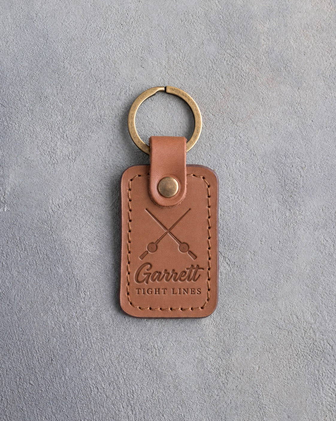 Caramel leather keychain debossed with crossed fishing rods above Garrett and TIGHT LINES, on gray plaster