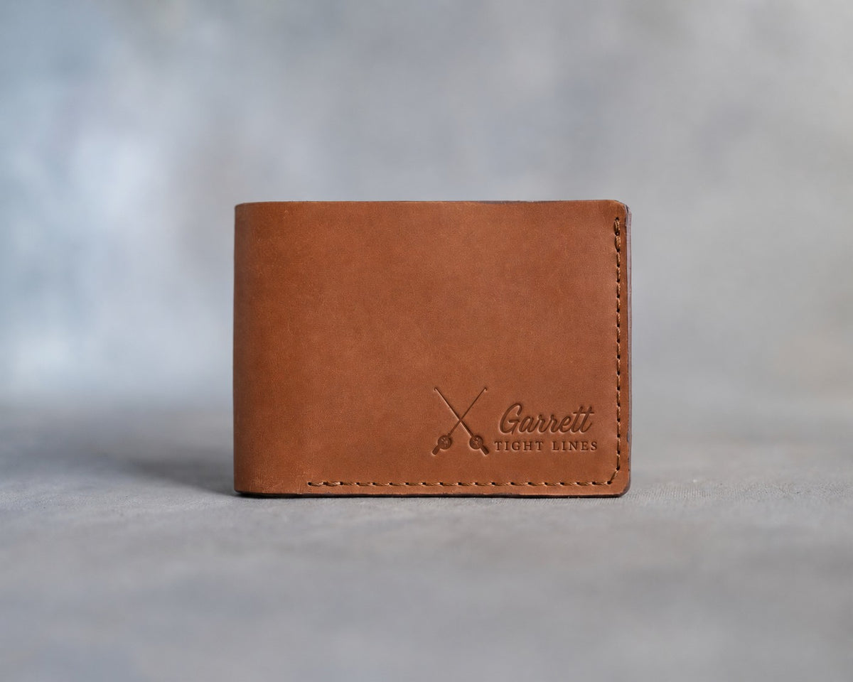 Closed caramel leather bifold wallet with crossed fishing rods beside Garrett and TIGHT LINES pressed in the lower right corner, on gray plaster