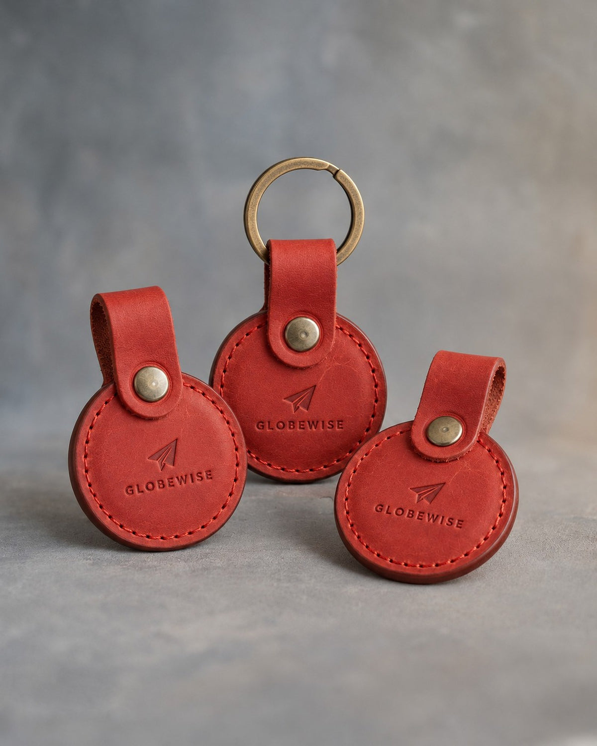 Poppy Red leather AirTag holder with snap loop and brass split ring, debossed with a sample business logo, on grey plaster