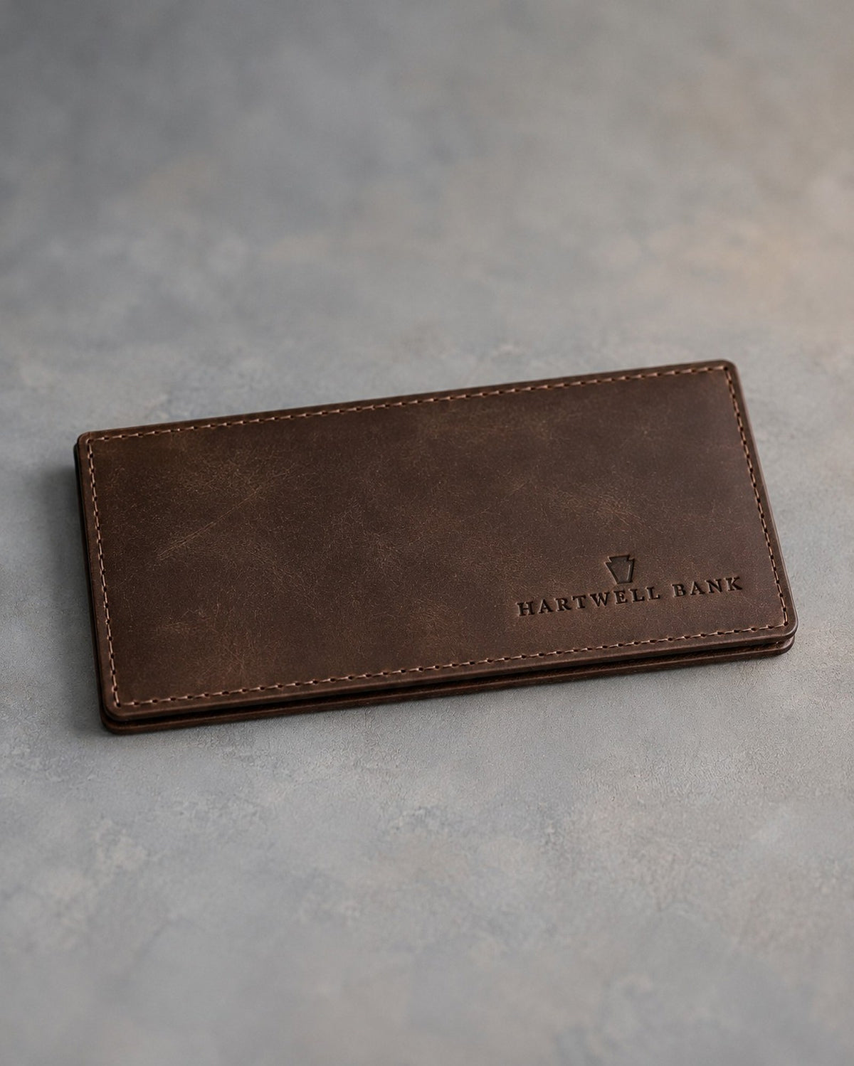 Dark Espresso leather checkbook cover debossed with a sample business logo, on a grey plaster backdrop