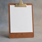 Caramel leather clipboard with brass clip, debossed with a sample business logo, on a grey plaster backdrop