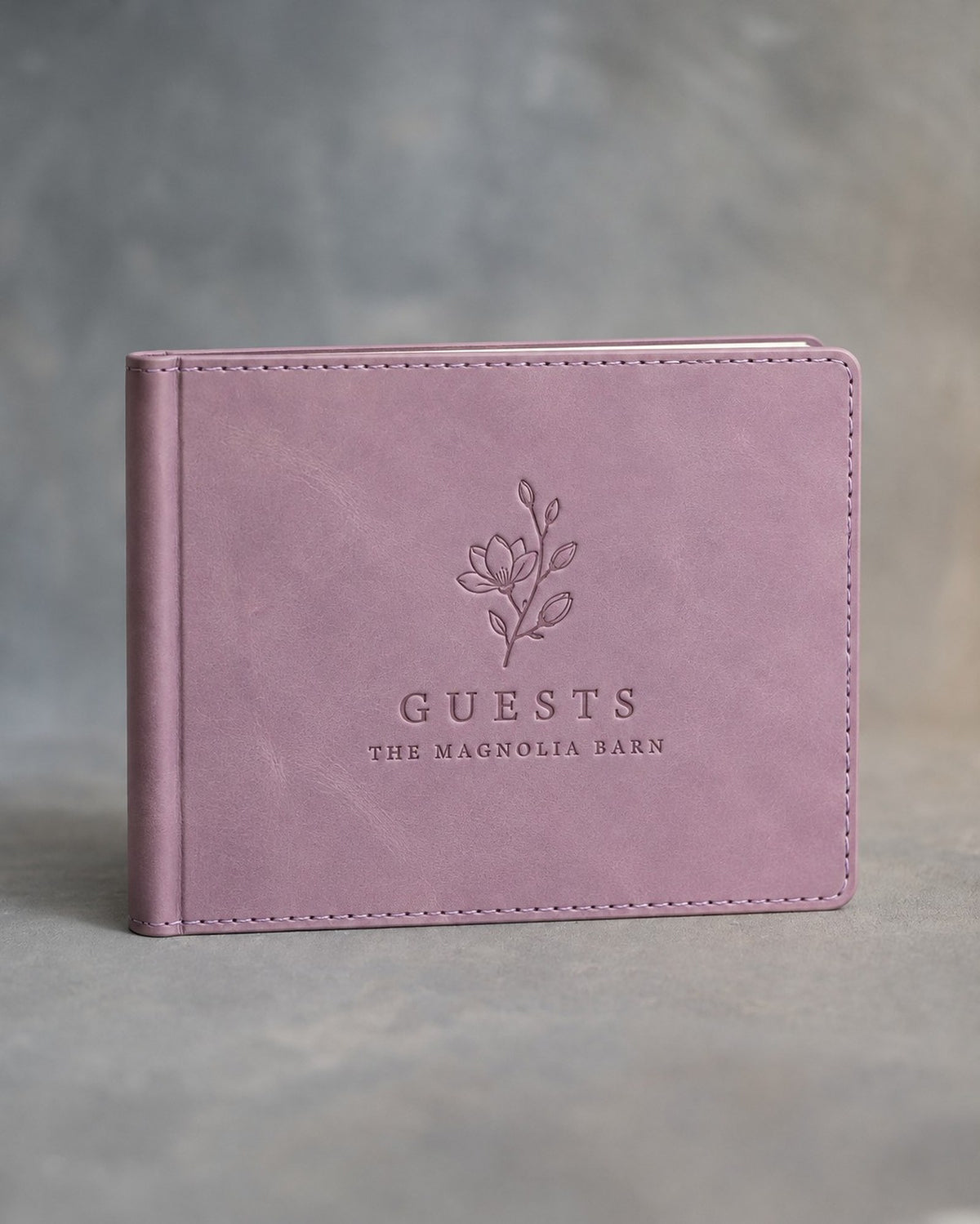 Lilac padded leather guest book in landscape format, debossed with a sample business logo, on a grey plaster backdrop