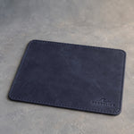 Blueberry leather mouse pad with stitched edge debossed with a sample business logo, on a grey plaster backdrop