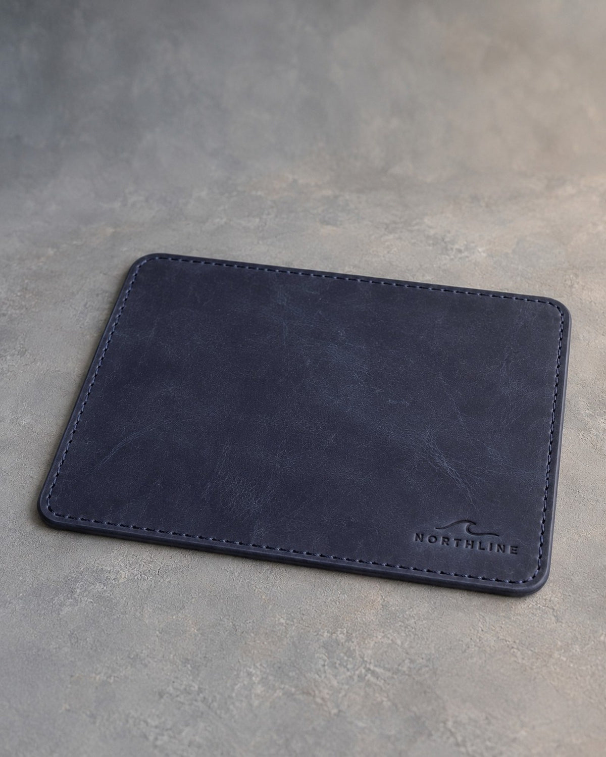Blueberry leather mouse pad with stitched edge debossed with a sample business logo, on a grey plaster backdrop