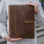 Sicilian Brown leather padfolio with snap strap closure and a sample logo debossed lower right, held in two hands