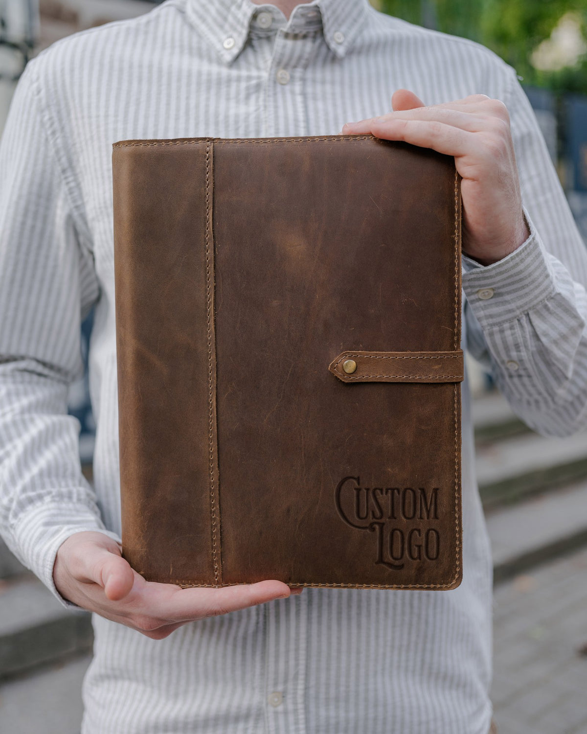Sicilian Brown leather padfolio with snap strap closure and a sample logo debossed lower right, held in two hands
