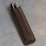 Dark Espresso leather pen case holding two pens, debossed lengthwise with a sample business logo, on a grey plaster backdrop