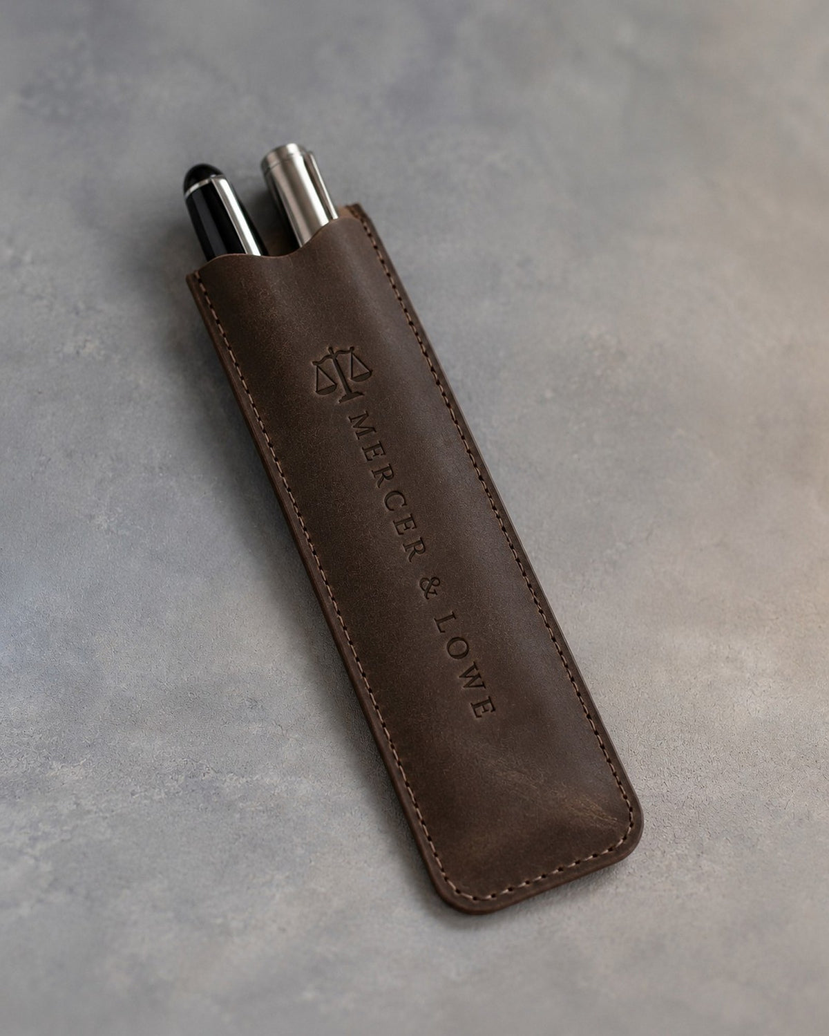 Dark Espresso leather pen case holding two pens, debossed lengthwise with a sample business logo, on a grey plaster backdrop