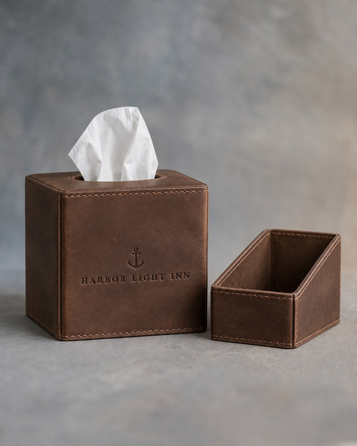 Sicilian Brown leather tissue box cover with oval top opening, debossed with a sample business logo, on grey plaster