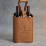 Caramel leather two-bottle wine tote with riveted handle, debossed with a sample business logo, on a grey plaster backdrop