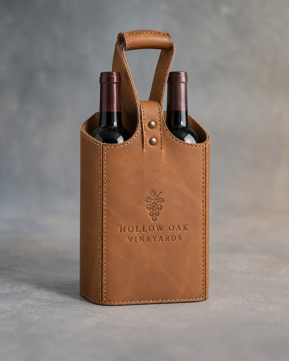 Caramel leather two-bottle wine tote with riveted handle, debossed with a sample business logo, on a grey plaster backdrop