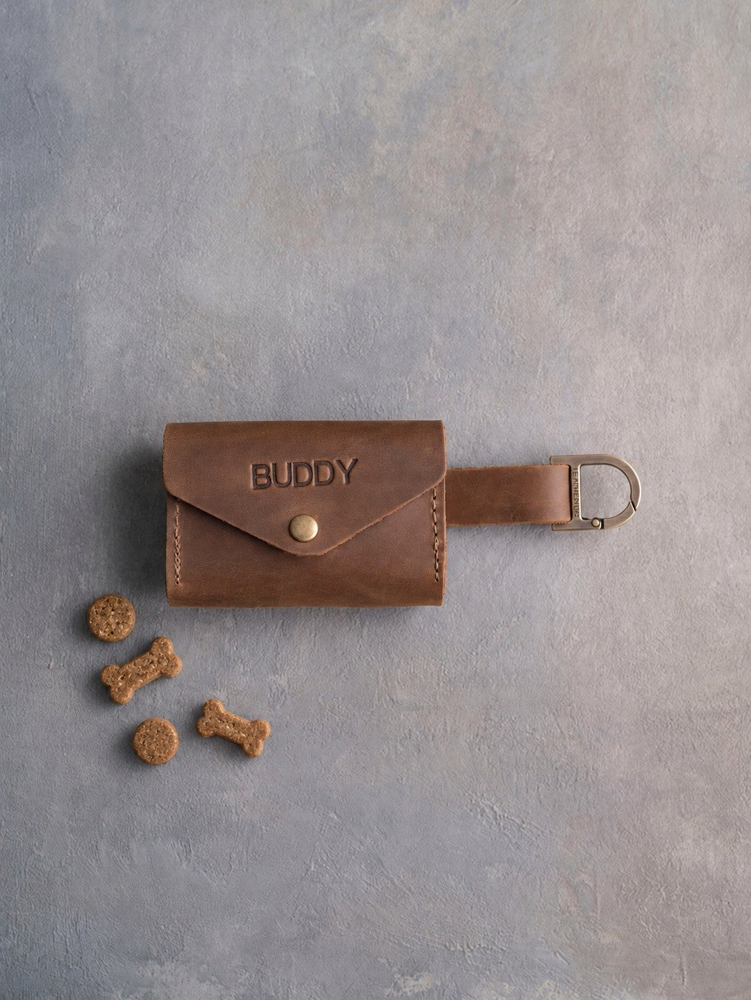 Sicilian Brown leather dog treat pouch with a snap flap stamped BUDDY, a strap with a D clip and small treats beside it