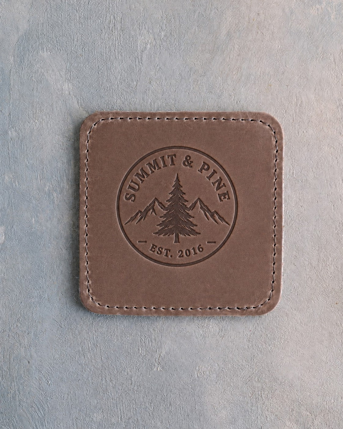 Sicilian Brown square leather coaster debossed with a sample round company logo, stitched edge, on grey plaster