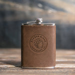 Sicilian Brown leather hip flask debossed with a sample round distillery logo, HARBOR & OAK DISTILLING CO., on a wooden table