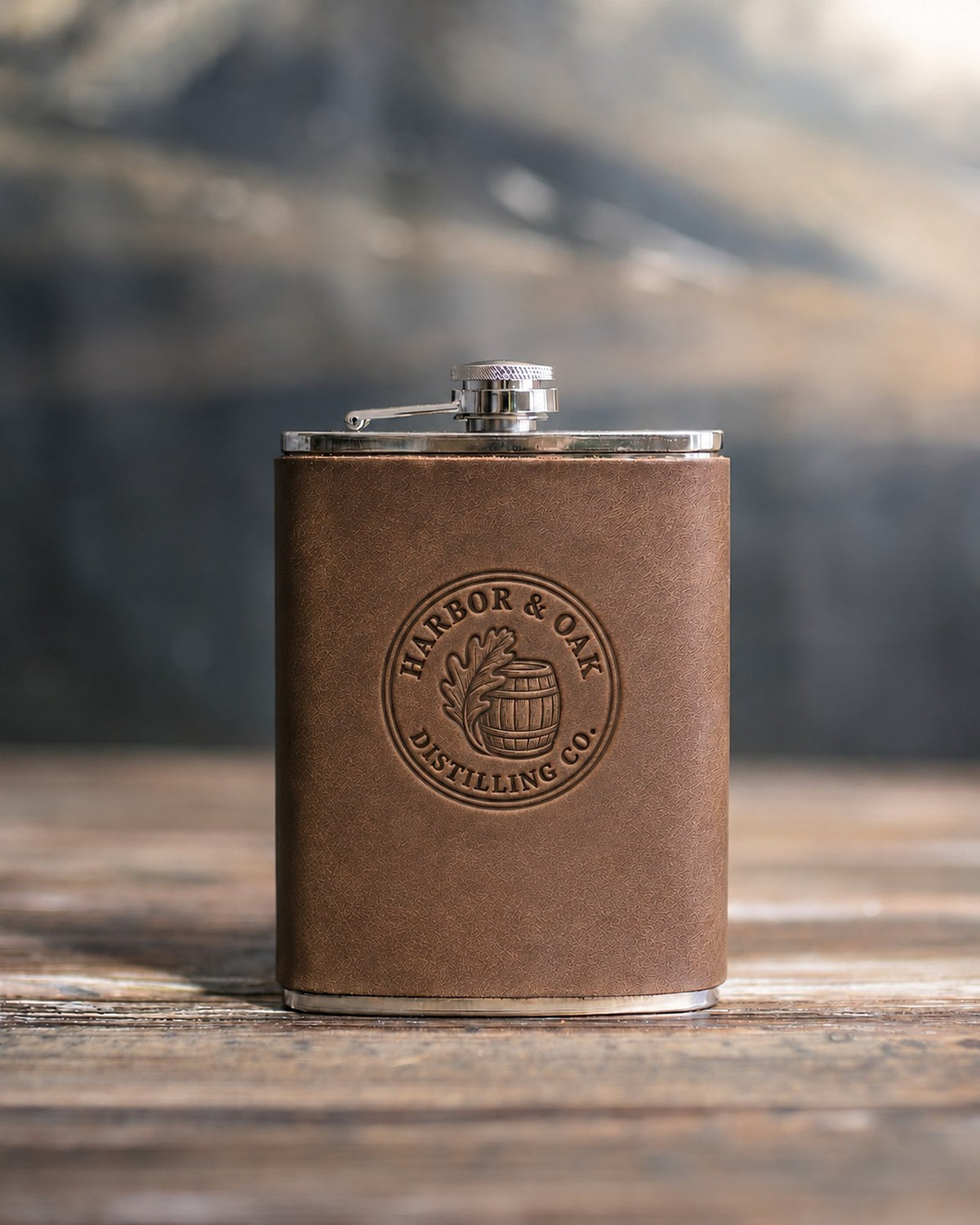 Sicilian Brown leather hip flask debossed with a sample round distillery logo, HARBOR & OAK DISTILLING CO., on a wooden table