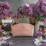 Blush leather makeup bag with a sample beauty studio logo, BELLE & BLOOM BEAUTY STUDIO, pressed into the front, among lilac flowers