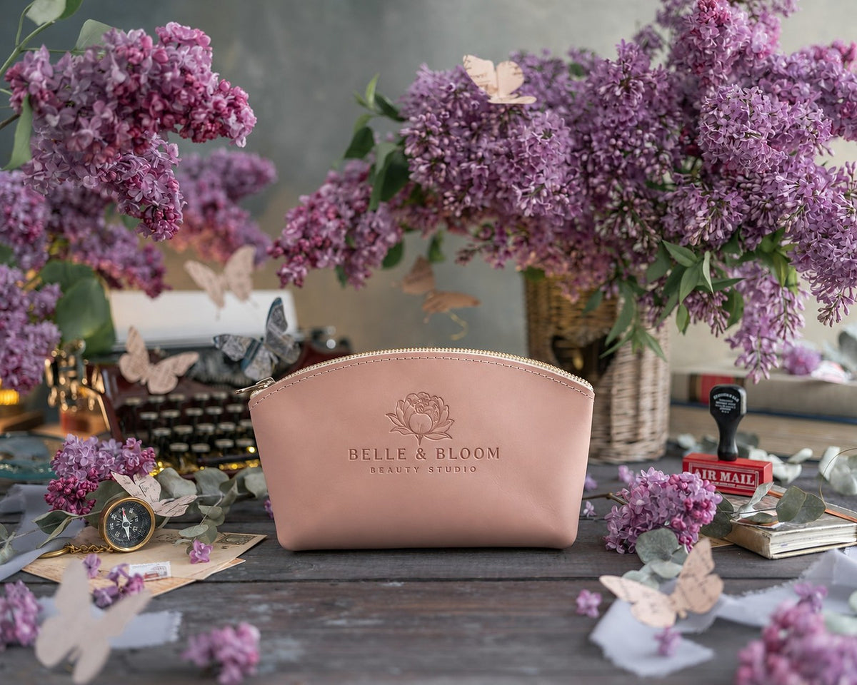 Blush leather makeup bag with a sample beauty studio logo, BELLE & BLOOM BEAUTY STUDIO, pressed into the front, among lilac flowers